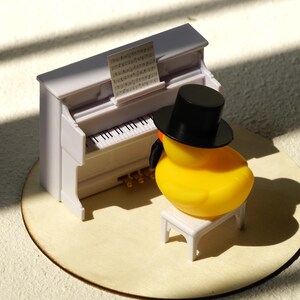 Peut inclure: Un canard en plastique jaune portant un chapeau haut de forme noir et un n&oelig;ud papillon noir est assis sur un petit tabouret blanc devant un piano blanc miniature. Une partition est pos&eacute;e sur le pupitre du piano. Les touches du piano sont noires et blanches.