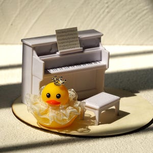 Peut inclure: Un piano droit miniature blanc avec une partition, un banc de piano blanc assorti et un canard en caoutchouc jaune portant une petite couronne argent&eacute;e et une collerette en dentelle blanche. L'ensemble est dispos&eacute; sur une base circulaire.