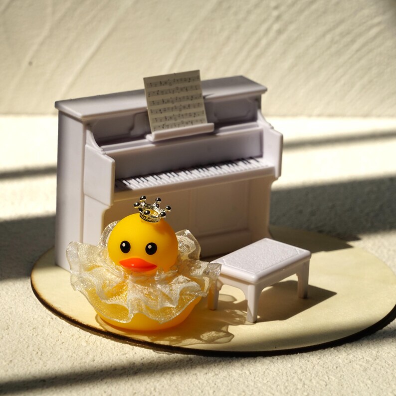 Peut inclure: Un piano droit miniature blanc avec une partition, un banc de piano blanc assorti et un canard en caoutchouc jaune portant une petite couronne argent&eacute;e et une collerette en dentelle blanche. L'ensemble est dispos&eacute; sur une base circulaire.