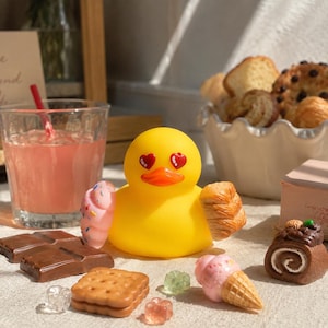 Puede incluir: Un pato de goma amarillo brillante con ojos rojos en forma de corazón y pico naranja. El pato sostiene una dona glaseada rosa en un ala y un croissant dorado en la otra, rodeado de barras de chocolate, galletas y helados en miniatura.