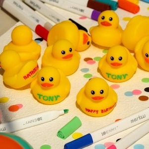 Könnte beinhalten: Eine Ansammlung gelber Gummi-Enten mit schwarzen Augen und orangefarbenen Schnäbeln. Einige Enten tragen den Aufdruck "TONY" oder "HAPPY BIRTH". Bunte Marker und Konfetti sind um die Enten herum verstreut.