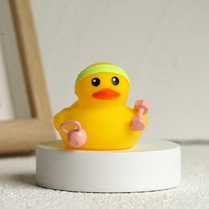 Peut inclure: Un canard en caoutchouc jaune portant un bandeau vert, tenant un haltère et une kettlebell roses. Le canard a des yeux noirs et un bec orange. Le canard est sur une plateforme circulaire blanche.