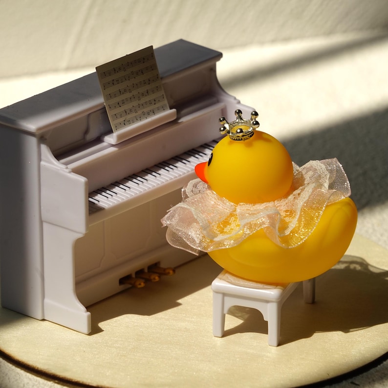 Peut inclure: Un canard en caoutchouc jaune portant une couronne argent&eacute;e et un col blanc &agrave; volants est assis sur un petit tabouret blanc devant un piano blanc miniature. Une partition musicale repose sur le pupitre du piano. Le canard a un bec orange.