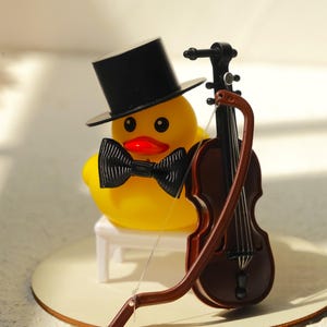 Könnte beinhalten: Eine gelbe Gummiente mit schwarzem Zylinder und Fliege, mit einer Miniatur-Geige und einem Bogen in Braun. Die Ente hat einen roten Schnabel und sitzt auf einem kleinen weißen Stuhl.