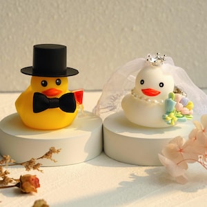 Könnte beinhalten: Zwei dekorative Gummienten für eine Hochzeit. Die gelbe Ente trägt einen schwarzen Zylinder und eine Fliege und hält ein kleines Glas. Die weiße Ente ist mit einem Schleier, einer Krone und einer Perlenkette geschmückt und hält einen Blumenstrauß.