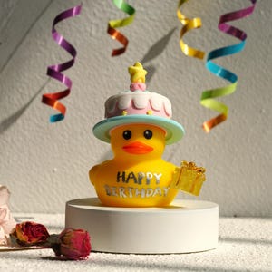 May include: A yellow rubber duck wearing a cake hat and holding a gift. The duck has the words "HAPPY BIRTHDAY" in silver lettering. Colorful streamers and dried roses are in the background.