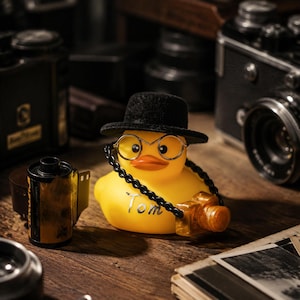 May include: A yellow rubber duck wearing a black fedora and thin-rimmed round glasses. A black chain hangs around its neck, holding a small orange camera. The name Tom is written in silver script on the front of the duck's chest.