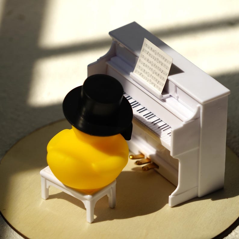 Peut inclure: Un canard en caoutchouc jaune portant un haut-de-forme noir est assis sur un petit banc blanc devant un piano droit miniature blanc. Le piano pr&eacute;sente des touches noires et blanches, trois p&eacute;dales dor&eacute;es et une partition sur le pupitre.