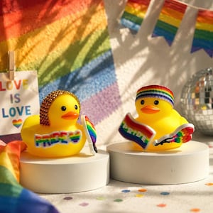 May include: Two yellow rubber ducks, each with a rainbow Pride flag and accessories. One duck has a gold-coloured hairpiece, the other a rainbow headband. The background includes a rainbow flag and the words "Love is Love".