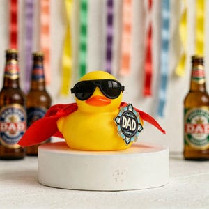 Pode incluir: Um pato de borracha amarelo usando óculos de sol pretos e uma capa vermelha, com um distintivo "DAD". O pato está em uma plataforma branca, com garrafas de cerveja no fundo e fitas coloridas.