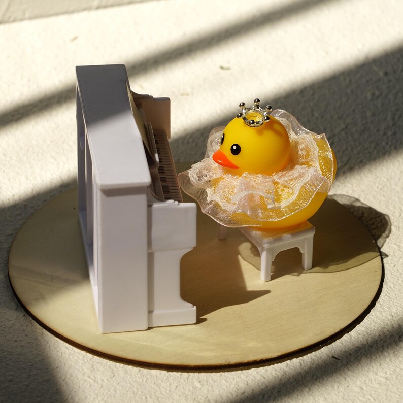 Peut inclure: Un canard en plastique jaune portant une couronne argent&eacute;e et un col &agrave; volants, assis devant un piano blanc miniature. Le canard est sur un petit banc blanc, pos&eacute; sur une surface en bois ronde et claire.