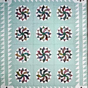 May include: A handmade quilt with a light teal background and a repeating pattern of pinwheel designs. The pinwheels are multicolored, and the quilt has a white border with a triangle pattern and floral accents.