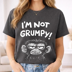 May include: Dark gray t-shirt with the text "I'M NOT GRUMPY!" in white. Below the text is a black and white illustration of a grumpy monkey face. The shirt has short sleeves and a crew neck.