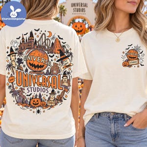 May include: An off-white t-shirt featuring a large, detailed graphic on the back and a smaller matching logo on the front left chest. The design includes orange and black illustrations of Universal Studios landmarks, pumpkins, ghosts, and bats.
