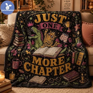 May include: A black fleece blanket featuring a colorful illustration of a green dinosaur and an orange cat reading an open book. The text reads JUST ONE MORE CHAPTER. Additional graphics include stacks of books, a trophy, glasses, and candles.