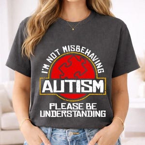 May include: Dark gray t-shirt with the text "I'M NOT MISBEHAVING AUTISM PLEASE BE UNDERSTANDING." The design features a red puzzle piece in a yellow and red circle. The shirt is a crew neck.