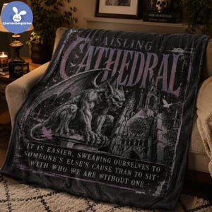 May include: A dark gray fleece blanket featuring a gothic cathedral illustration, a gargoyle perched on a ledge, and the text AISLING CATHEDRAL. Below the image, the text reads: IT IS EASIER, SWEARING OURSELVES TO SOMEONE'S ELSE'S CAUSE THAN TO SIT WITH WHO WE ARE WITHOUT ONE.