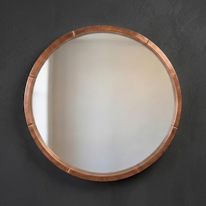Puede incluir: Un espejo redondo con marco color cobre. El marco tiene una serie de detalles rectangulares en el borde. El espejo refleja una pared de color claro. Un artículo decorativo.