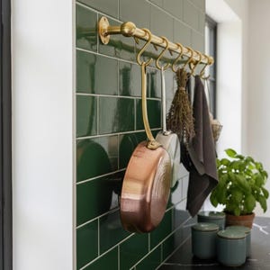 Könnte beinhalten: Eine Küchenszene mit einer Messing-Topfhalterung, an der Kochutensilien hängen, vor einer dunkelgrünen gefliesten Wand. Eine Kupferpfanne und andere Küchenutensilien hängen an der Halterung. Eine Topfpflanze und kleine tealfarbene Behälter stehen auf der Arbeitsplatte.