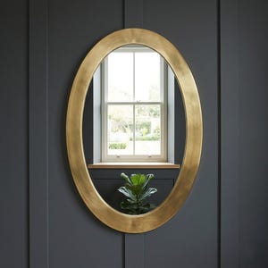 May include: An oval-shaped mirror with a gold-colored frame hangs on a dark gray wall. The mirror reflects a window with a view of a garden and a small plant. The frame has a smooth, metallic finish.