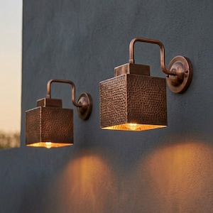 May include: Two copper-colored outdoor wall sconces. Each sconce has a square, textured shade and a curved arm. The sconces are mounted on a dark gray wall. The light from the bulbs creates a warm glow.