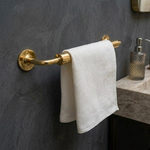 May include: A brass-coloured towel rail fixed to a dark grey wall, holding a folded white towel. The rail has a curved design with circular mounting plates. The towel is made of a textured fabric.