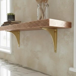 May include: A copper-finished floating shelf with a hammered texture, supported by two gold-colored brackets. The shelf is mounted on a light-colored wall, and a window is visible in the background. A small potted plant and books are on the shelf.