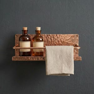 Puede incluir: Estante de pared de color cobre con dos botellas de vidrio marrón con tapones de corcho y etiquetas en blanco. Una toalla beige cuelga debajo. El estante tiene una textura martillada y una barra para la toalla.