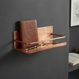 May include: A copper-colored wall shelf with a rectangular base and a curved front rail. A brown leather journal, a pair of glasses, and a black pen rest on the shelf. The shelf is mounted on a dark gray wall.