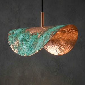 May include: A modern pendant light fixture with a unique, curved design. The light features a copper finish with a textured, hammered surface. One side of the fixture has a green patina, adding a contrast to the copper. The light hangs from a thin black cord.
