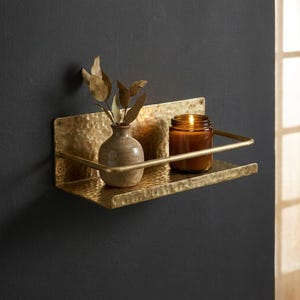 May include: A hammered brass wall shelf with a small vase holding dried leaves and a lit candle in a glass jar. The shelf has a curved metal bar to keep items from falling.
