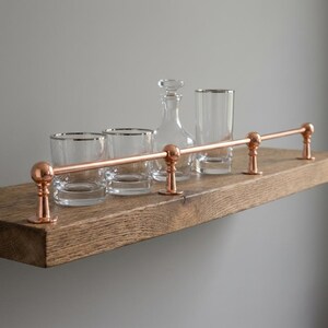 May include: A wooden shelf with copper-colored hardware, holding clear glassware and a glass decanter. The shelf has a copper bar with decorative ball ends. The glassware includes two lowball glasses and two taller glasses with silver rims.