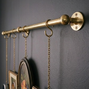 May include: A brass-colored wall-mounted picture rail with decorative hooks and chains. Several framed pictures are suspended from the rail. The rail is mounted on a dark gray wall, creating a vintage aesthetic.