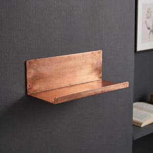 May include: A rectangular wall-mounted shelf crafted from polished copper with a warm metallic finish. The shelf features a flat horizontal surface with a small front lip and a tall vertical back panel secured by four visible screws at the corners.