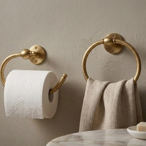Op de afbeelding: Een goudkleurige badkamerset met een toiletpapierhouder met een rol wit papier en een handdoekring met een beige handdoek. De set is gemonteerd op een neutraal gekleurde muur.