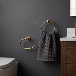 May include: A bathroom scene featuring a dark gray waffle-weave towel hanging from a brass towel ring. A matching brass towel holder is mounted on the wall. The fixtures are set against a dark gray wall, with a wooden vanity and a white countertop.