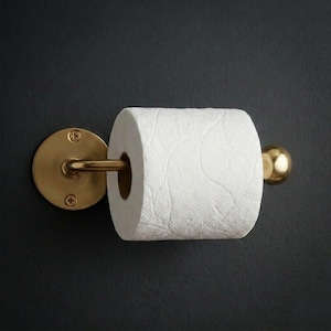Puede incluir: Un portarrollos de papel higiénico de latón con un rollo completo de papel higiénico blanco. El soporte tiene una placa trasera redonda y un brazo curvo con un extremo esférico. El accesorio está montado sobre un fondo oscuro.