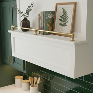May include: A white cabinet with a brass bar and decorative accents. A framed fern print, books, and a dark vase with greenery sit on the cabinet. The cabinet is above a green tiled backsplash.