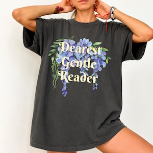 May include: A charcoal gray short-sleeve t-shirt featuring a graphic of blue wisteria flowers and the text "Dearest Gentle Reader" printed in a cream-colored serif font. The shirt has a crew neckline and a relaxed, loose-fitting silhouette.