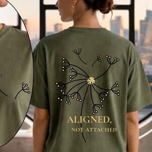 May include: Olive green t-shirt featuring a black dandelion design on the back with the words "ALIGNED, NOT ATTACHED" in gold. The front of the shirt has a smaller dandelion graphic. The shirt is made of a soft material.
