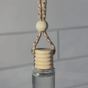 May include: A small, clear glass bottle filled with liquid, suspended by a beige rope with gold detailing. The bottle has a light wood cap. The background is a white brick wall.