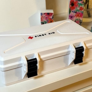 Könnte beinhalten: Eine weiße, rechteckige Erste-Hilfe-Box mit schwarzen Verschlüssen und dem Schriftzug "FIRST AID" in Schwarz. Ein rotes Kreuz befindet sich oben. Die Box steht auf einer weißen Oberfläche.
