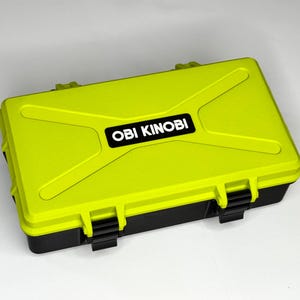 以下が含まれることがあります： 蓋に「OBI KINOOBI」の文字が入った、ライムグリーンと黒の長方形のケース。ケースはテクスチャ加工された表面と黒い留め金が付いています。ケースは白い表面にあります。