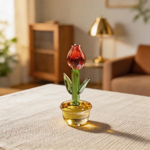 May include: A glass tulip ornament featuring a red flower, green stem and leaves, and a yellow pot. The ornament is placed on a beige tablecloth. The background includes a wooden cabinet, a gold lamp, and a brown armchair.
