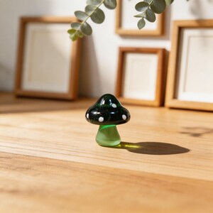 May include: A small, decorative glass mushroom with a dark green cap and white polka dots. The stem is a lighter green color. The mushroom is sitting on a wooden surface.