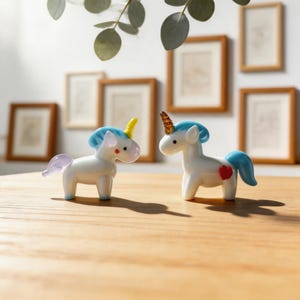 May include: Two white unicorn figurines with blue manes and tails, and colorful horns. One has a yellow horn and the other has a brown horn. One has a red heart on its side. The figurines are on a wooden surface.
