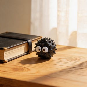 May include: A black, spiky ball with large white eyes and a black leather-bound journal with a black strap sit on a wooden surface. The ball has a cartoonish appearance. The journal is closed. Sunlight streams in from a sheer curtain in the background.