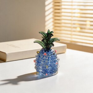 Può includere: Una scultura decorativa a forma di ananas in vetro. L'ananas è blu con un design a spirale, foglie verdi e piccoli accenti rossi e dorati. È appoggiata su una superficie bianca vicino a un libro chiuso.