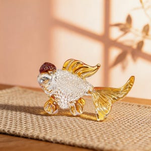 May include: A decorative glass goldfish figurine with a clear body, gold fins, and a brown head. The fish is posed on a woven surface, showcasing its intricate details and craftsmanship. A unique home decor item.