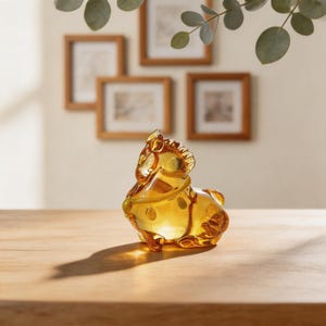 May include: A decorative glass horse figurine in a warm amber color. The horse is posed in a seated position with detailed features, including a mane and tail. The figurine is on a wooden surface.